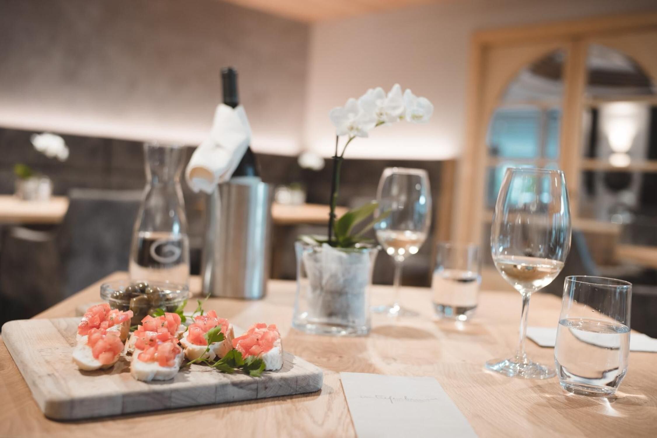 A dining table with a wooden surface, a marble serving board with appetizers, a vase with white flowers, a bottle, a glass pitcher, and two glasses of white wine.