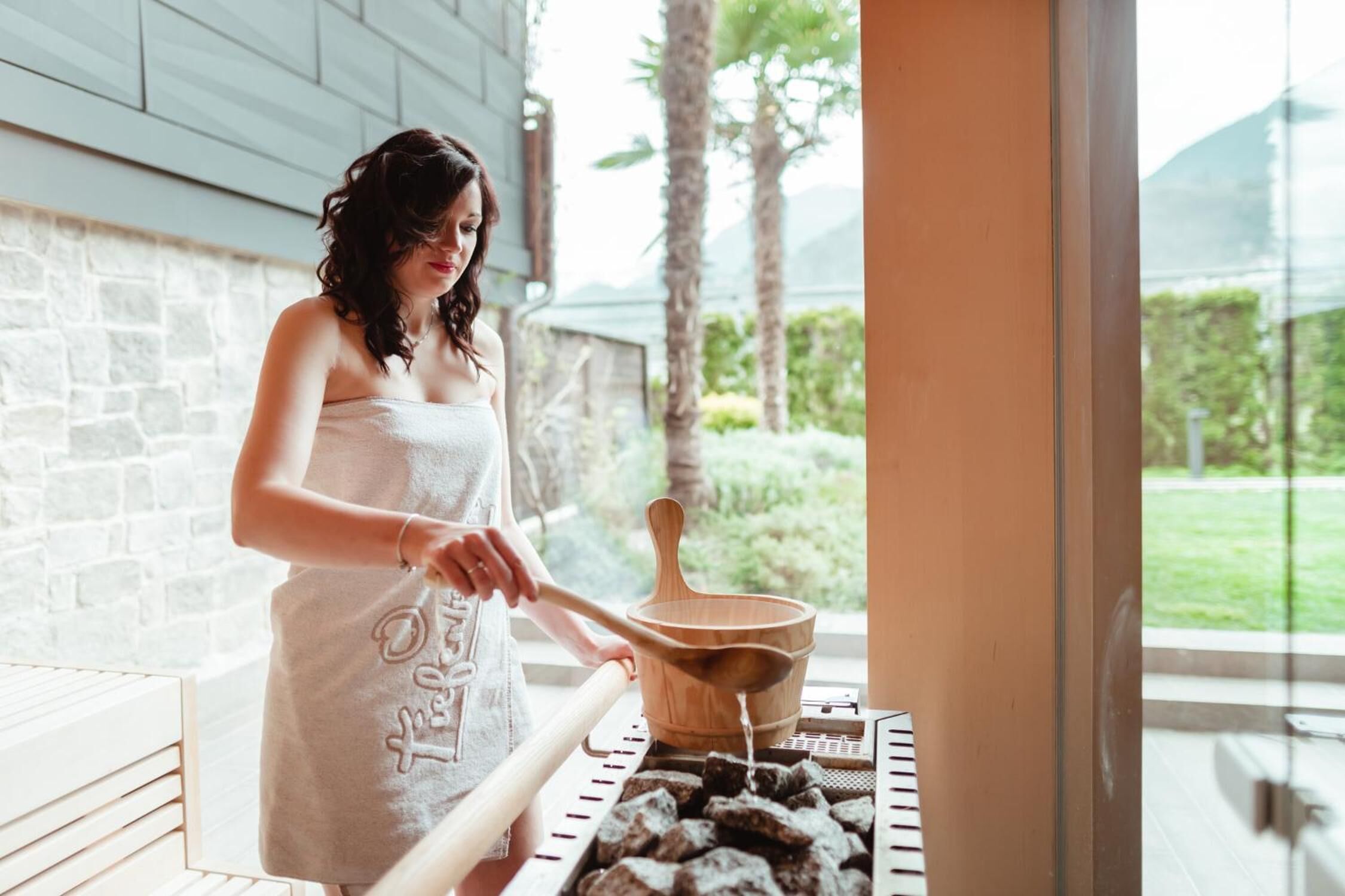 A person in a towel near a grill with charcoal, outdoors with a stone wall and greenery in the background.
