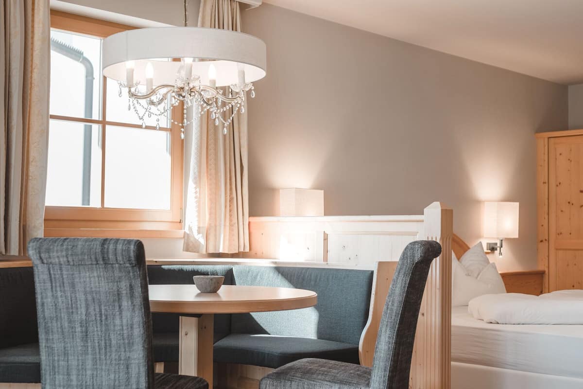 A hotel room with a round dining table, two upholstered chairs, a chandelier, a bed with white bedding, and a wooden wardrobe.