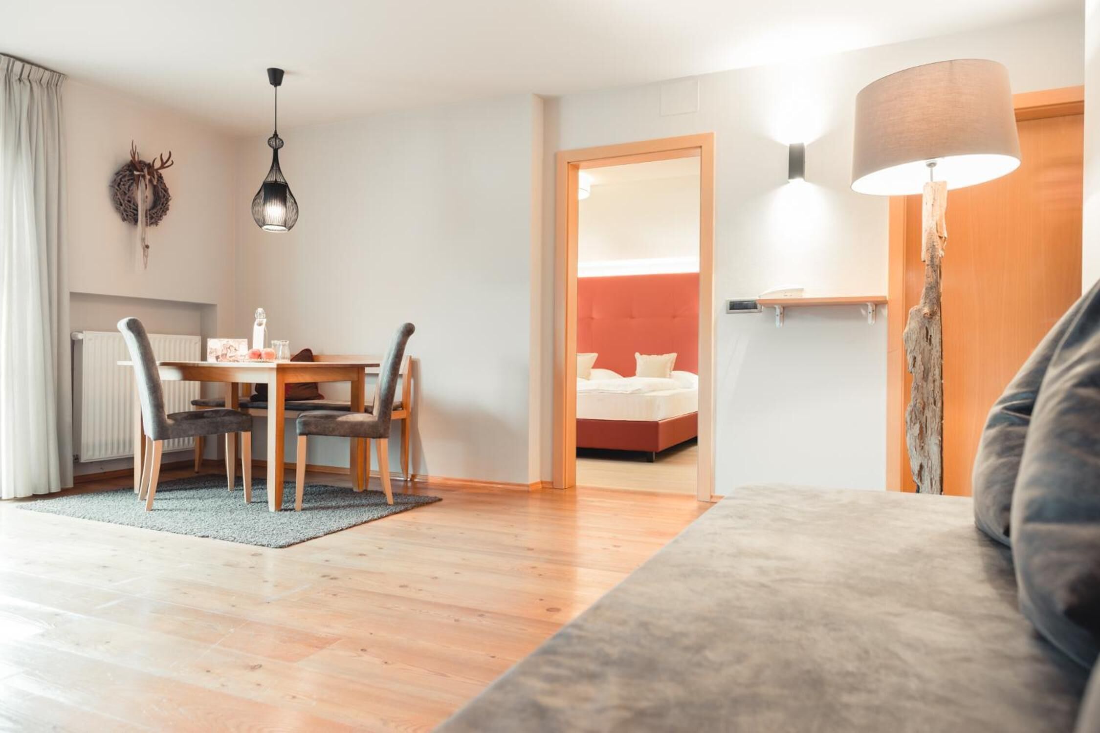 Una habitación de hotel moderna con una mesa de comedor de madera, dos sillas, un sofá, una lámpara y una cama con cabecera roja.