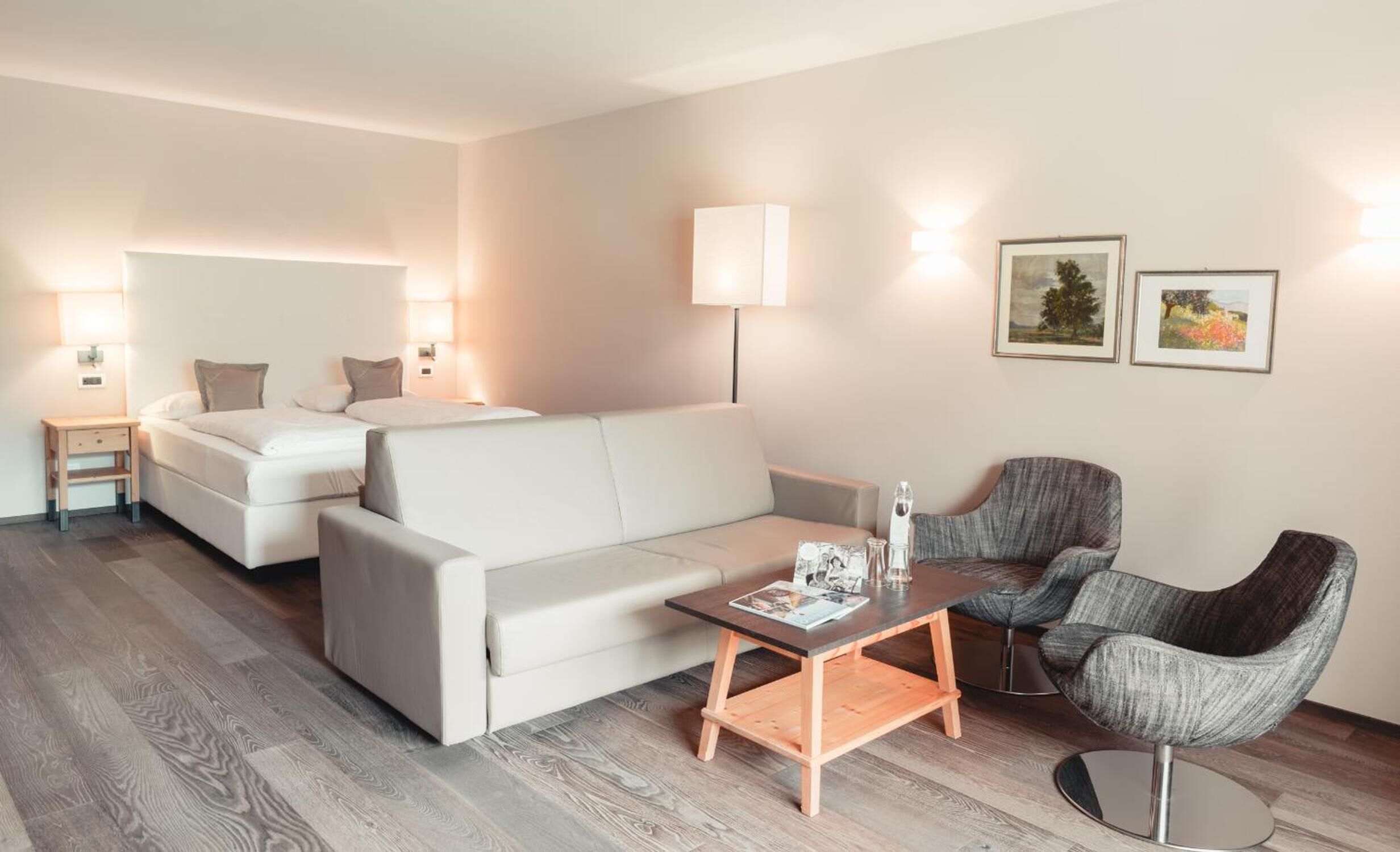 Ein modernes Hotelzimmer mit einem Bett, einer Couch, einem kleinen Tisch und einem Sessel. Zwei Wandlampen und zwei gerahmte Bilder sind an der Wand angebracht.