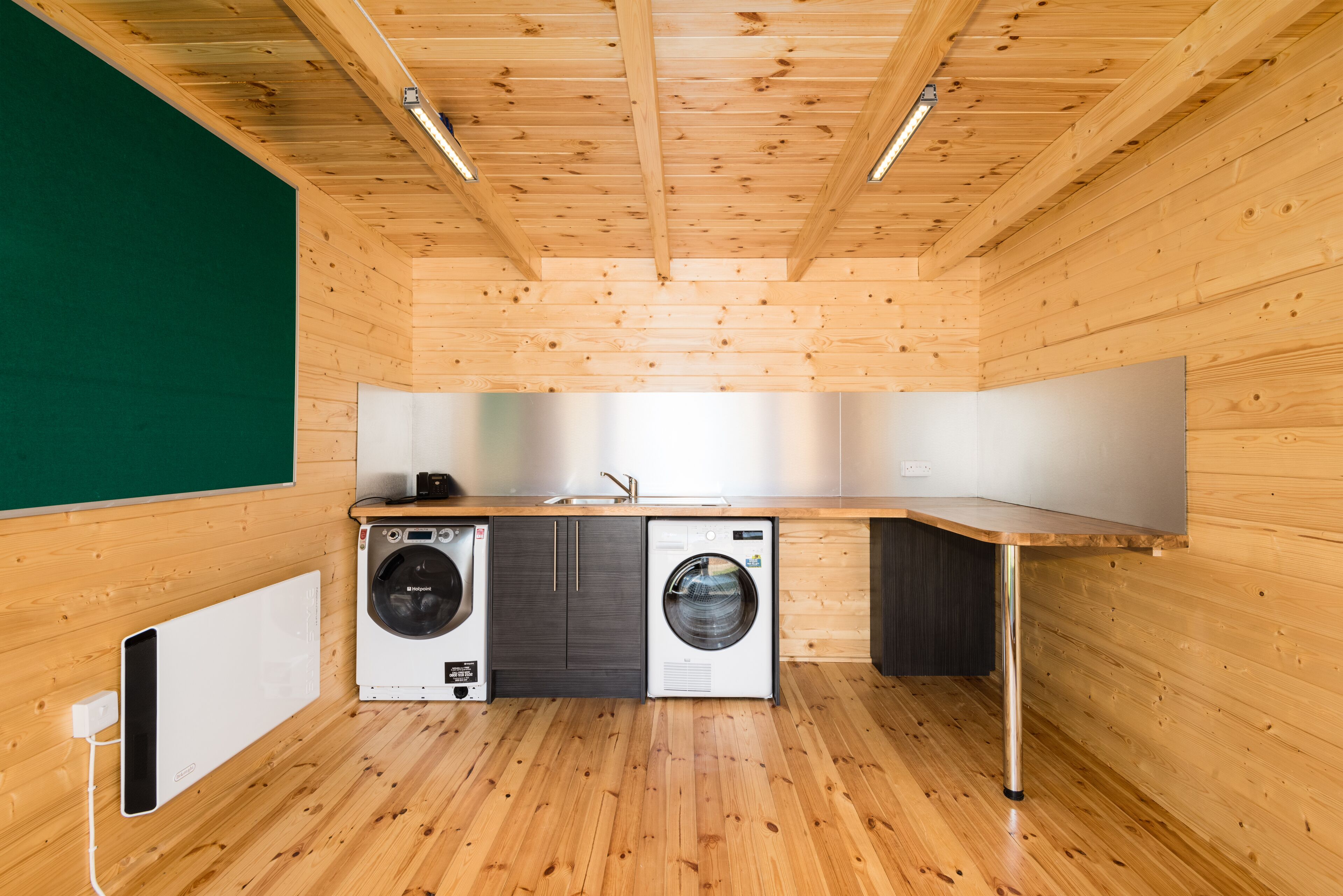 A laundry room with wooden walls and floors, two washing machines, a dryer, and a countertop with a sink.