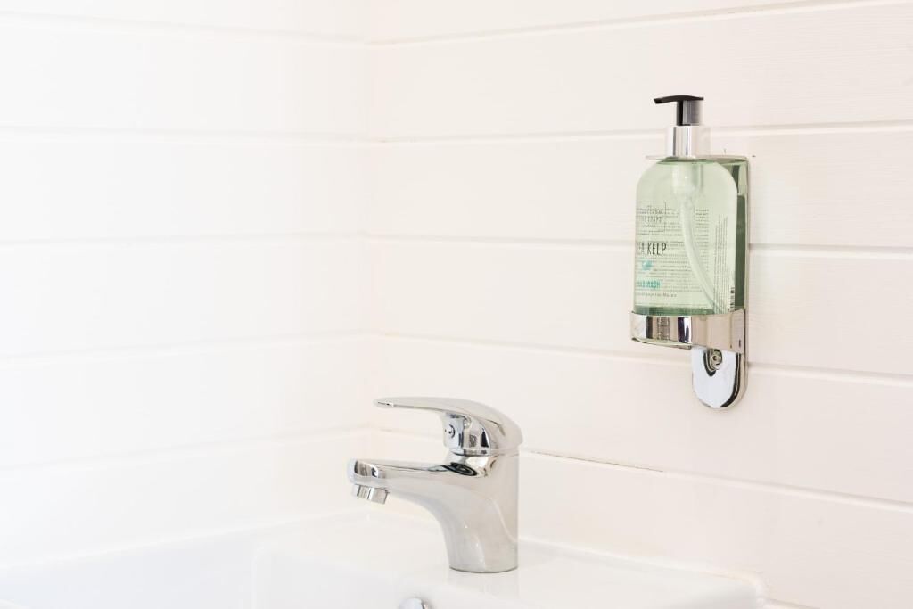A bathroom with a white tiled wall, a chrome faucet, and a soap dispenser mounted on the wall.
