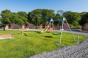 A playground with swings and a slide, surrounded by wooden structures with windows. - Weedingshall Lodges (Falkirk)