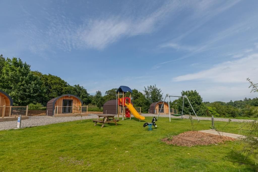 A playground with a slide and swings, surrounded by wooden cabins with a picnic table nearby.