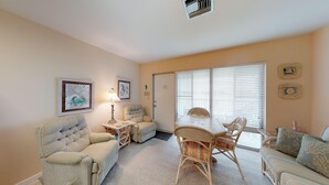 Una sala de estar acogedora con sofá, sillón, mesa de comedor y sillas. Hay cuadros en la pared, una lámpara en la mesita auxiliar y una puerta con ventanal.
