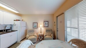 Cocina compacta con armarios blancos, refrigerador y pileta. Hay dos sillones y una pequeña mesa con una lámpara. La habitación tiene un ventanal con persianas y una puerta.