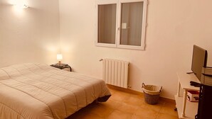 Een slaapkamer met een bed, nachtkastje, lamp, radiator, raam, televisie en een klein tafeltje met een mandje.