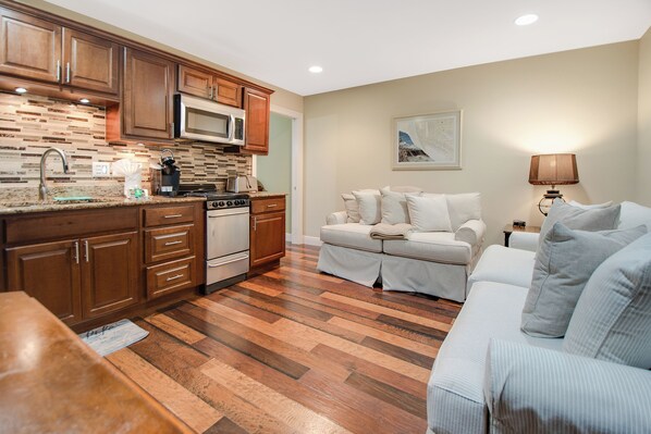 A modern kitchen with wooden cabinets, a microwave, and a stove. There is a dining area with a table and chairs, and a living room with a sofa and a lamp.