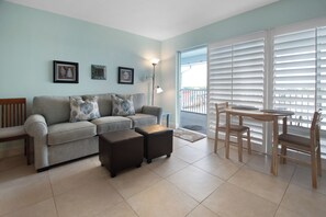 A living room with a sofa, two ottomans, a dining table with chairs, and a balcony with white shutters.