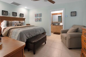 Quarto com cama, um banquinho, cômoda, cadeira e banheiro com pia e espelho.