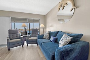 A living room with a blue sofa, two armchairs, a round mirror, and a view through large windows.