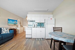 A compact kitchen with white cabinets, a dining table with a lace tablecloth, a blue sofa, and a painting of a beach scene.