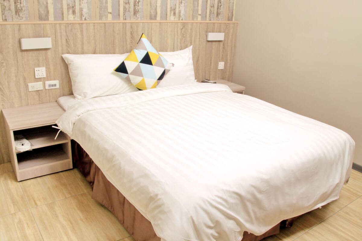 A neatly made bed with a white comforter and a geometric-patterned pillow. There is a wooden nightstand with a drawer and a small shelf. The room has a wooden headboard and a plain wall with a light switch and a power outlet.