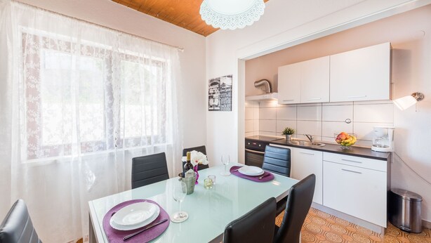 Un coin repas avec une table dressée pour deux personnes, une cuisine avec des meubles blancs et un luminaire au plafond.