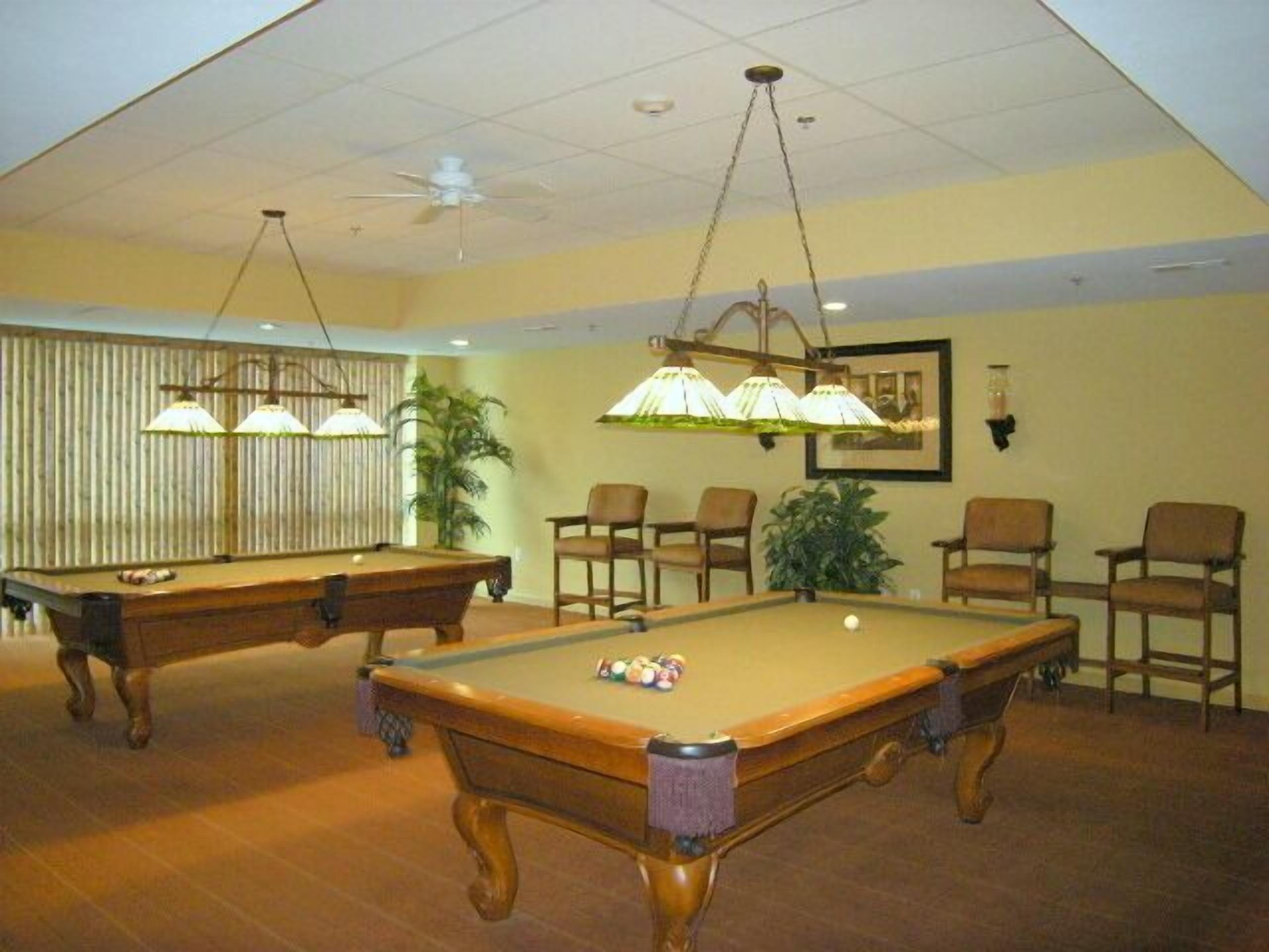 A pool table with balls, a ceiling fan, and several chairs in a room with wooden flooring and a yellow wall.