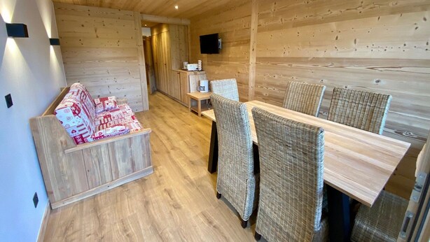 Een gezellige kamer met een houten plafond, een bank met gestreepte kussens, een eettafel met rieten stoelen en een flatscreen televisie aan de muur.