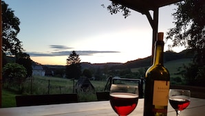 Ein Tisch mit einer Weinflasche und zwei Gläsern, mit Blick auf eine malerische Landschaft: ein Sonnenuntergang über einem Tal mit Häusern und Bäumen.