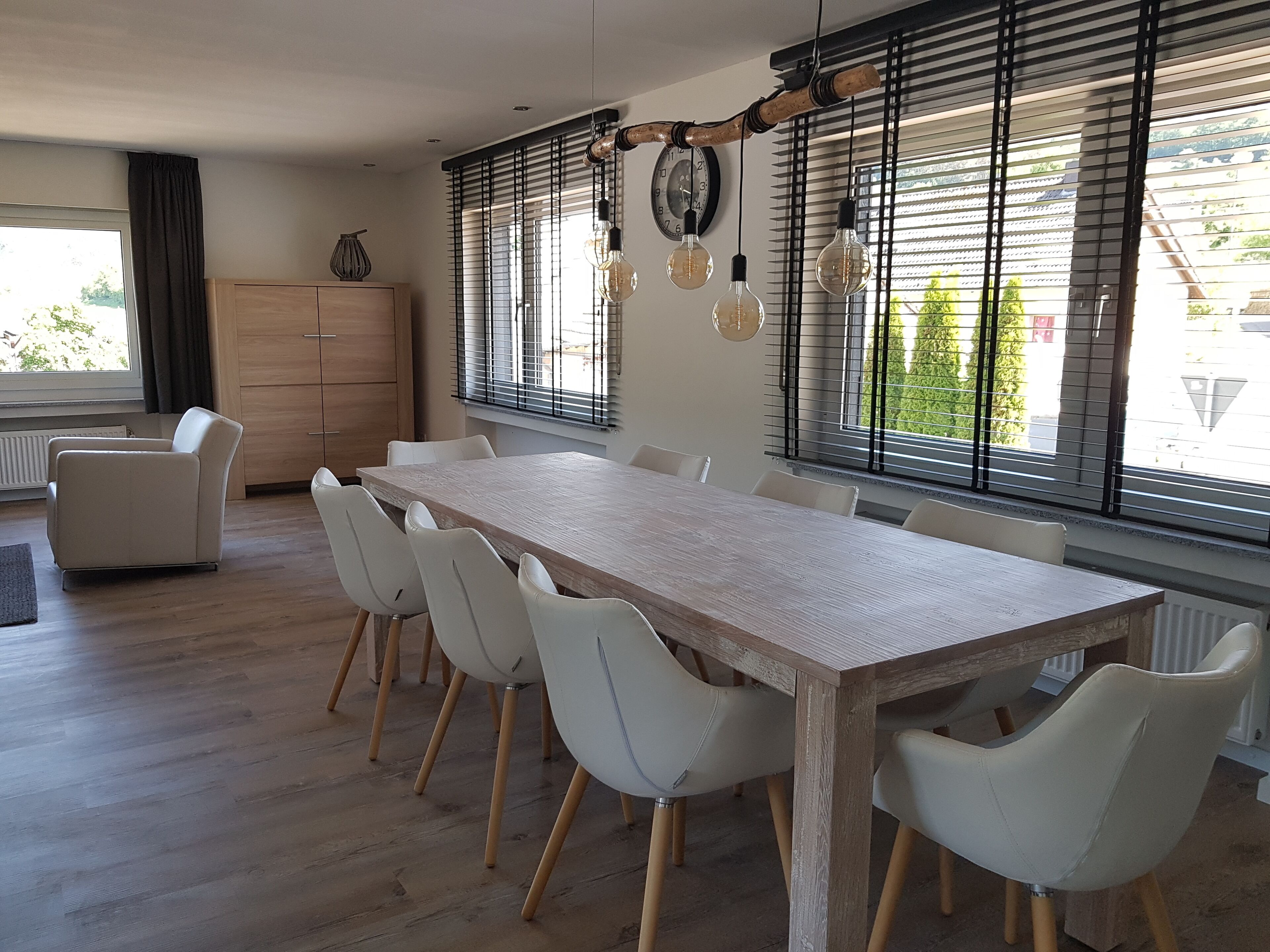 A modern dining area with a long wooden table and white chairs, large windows with blinds, and hanging light bulbs.