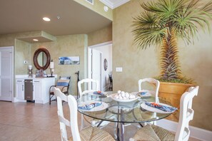 A dining area with a glass table surrounded by white chairs, a large potted plant, and a wall-mounted mirror.