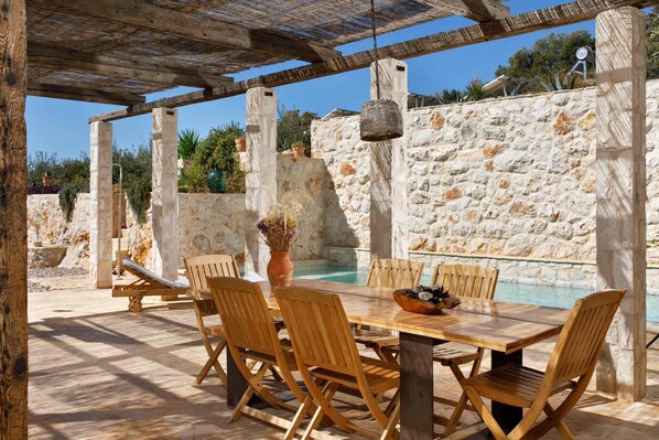 A wooden table and chairs set up outdoors with a stone wall and a pool in the background.