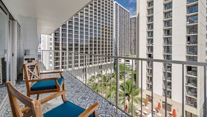 A balcony with two wooden chairs and a small table, overlooking a cityscape with tall buildings and palm trees. - Gorgeous 2-bedroom listing! Half-block from Waikiki Beach! (Honolulu)