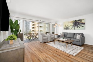 A modern living room with a grey sofa, two armchairs, a wooden coffee table, and a large window overlooking a cityscape. - Gorgeous 2-bedroom listing! Half-block from Waikiki Beach! (Honolulu)