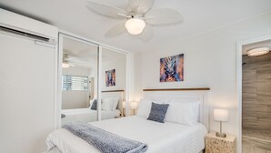 A bedroom with a large bed, a ceiling fan, a mirror, and artwork on the wall. - Gorgeous 2-bedroom listing! Half-block from Waikiki Beach! (Honolulu)