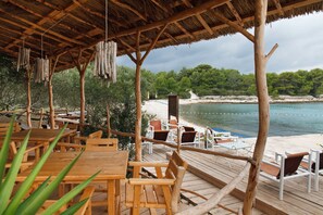 A wooden deck with tables and chairs overlooking a beach and water. - Robinson House Nicolina (Hvar)