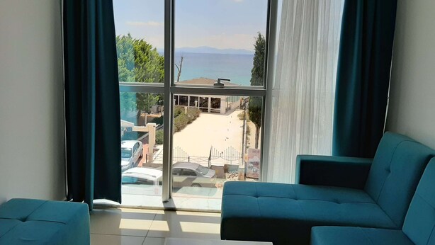 Habitació amb una finestra gran que ofereix vistes a una platja i al mar. Hi ha dos sofàs blaus i una taula petita a la habitació.