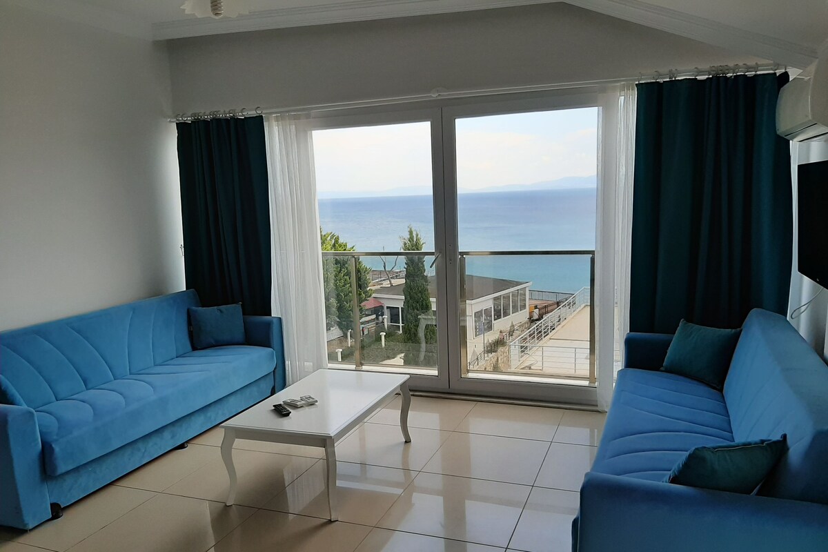 A living room with two blue sofas, a white coffee table, and a large sliding glass door leading to a balcony with a view of the sea.