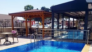 Una piscina rectangular d'aigua cristal·lina blava, envoltada per un solàrium de fusta i una zona coberta amb taules i cadires.