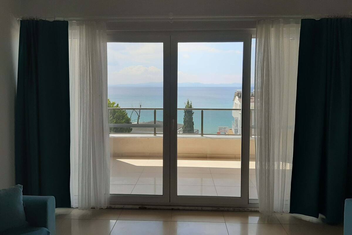 A room with a large sliding glass door leading to a balcony with a view of the sea and distant mountains.