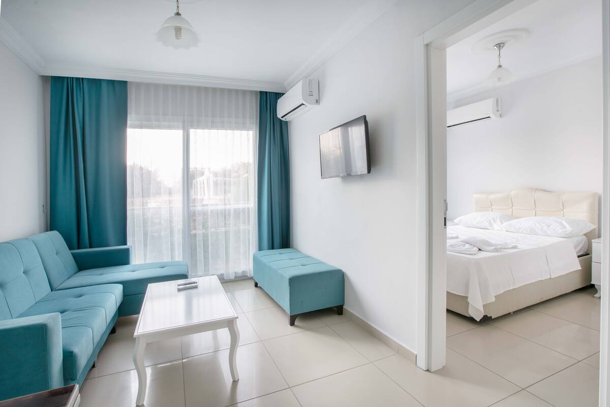 A modern hotel room with a sofa, ottoman, bed, and a flat-screen TV mounted on the wall.