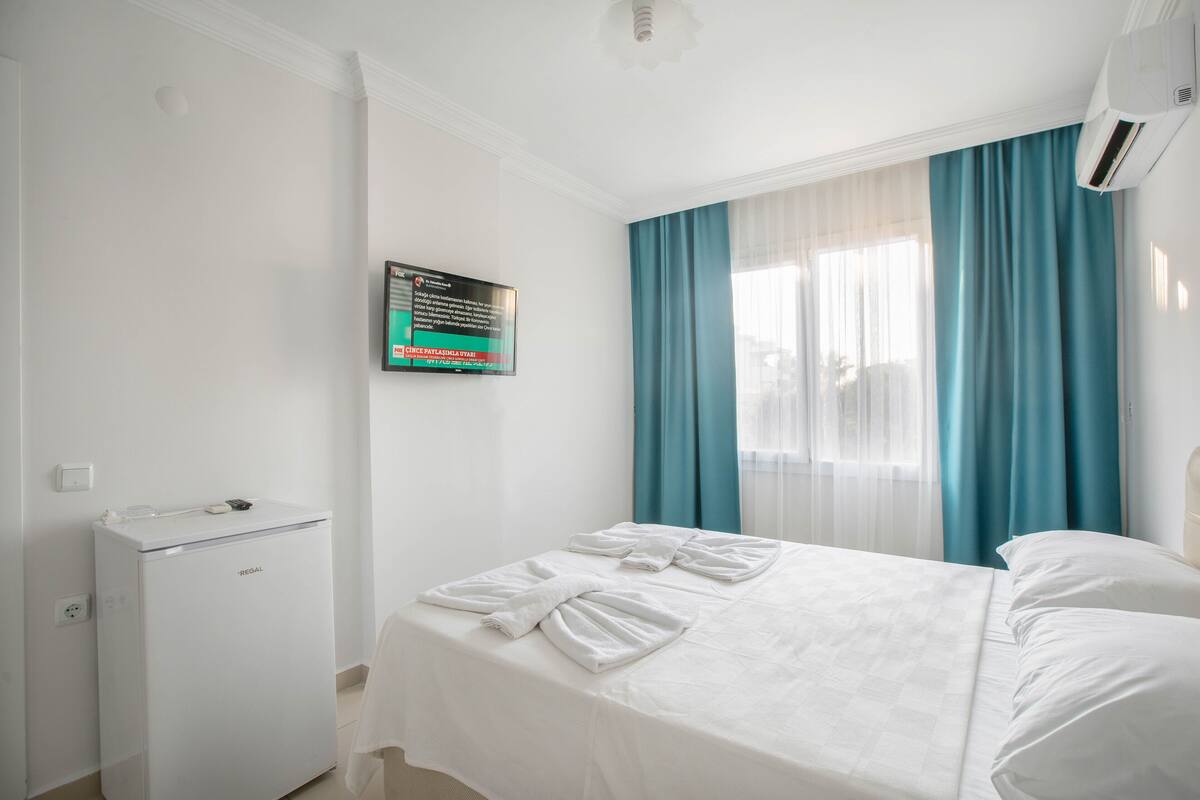 A hotel room with a bed, a small refrigerator, a wall-mounted TV, and a window with blue curtains.