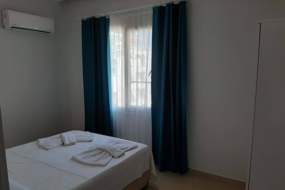 A hotel room with a bed, a window with curtains, an air conditioning unit, and a wardrobe.