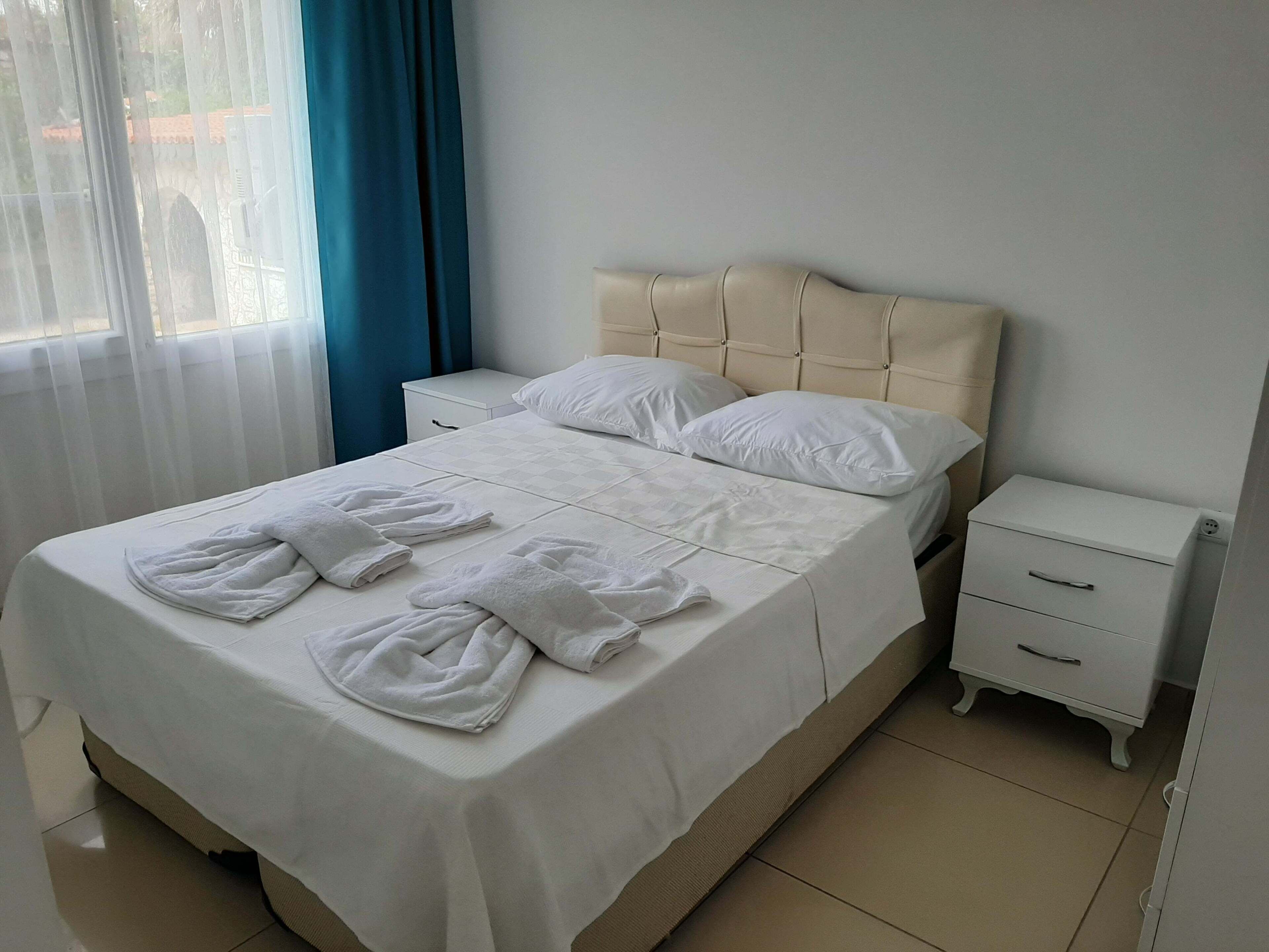 Un lit bien fait, avec du linge de lit et des serviettes blanches, une table de chevet dotée d’un tiroir et une fenêtre ornée de rideaux bleus.