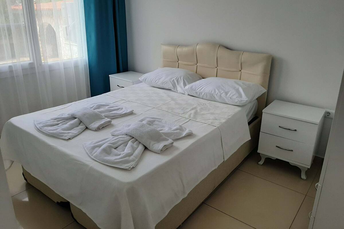 A neatly made bed with white linens and towels, a bedside table with a drawer, and a window with blue curtains.