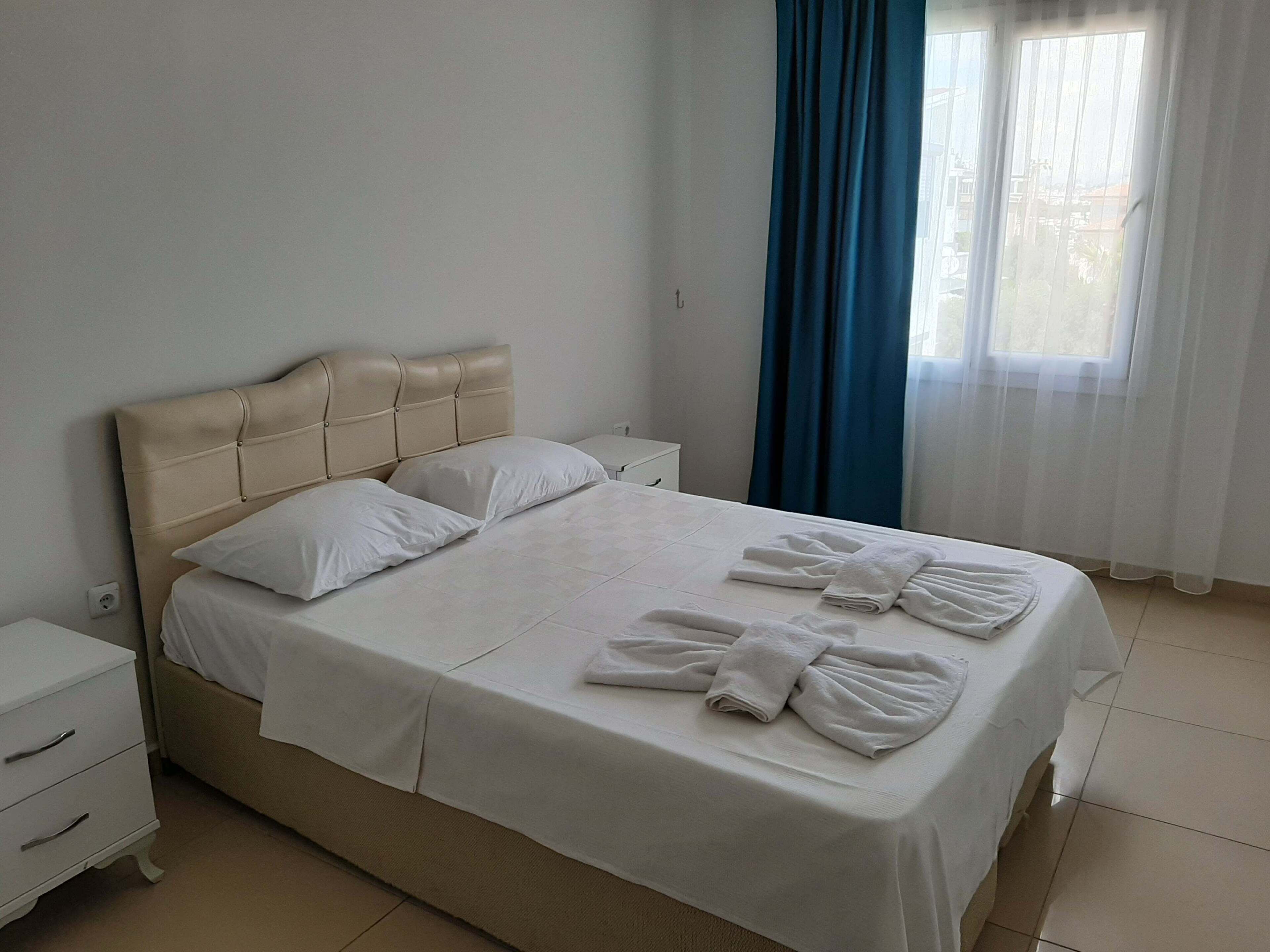 Un lit bien fait, avec du linge de lit et des serviettes blanches, une tête de lit beige, une table de chevet blanche et une fenêtre ornée de rideaux bleus.