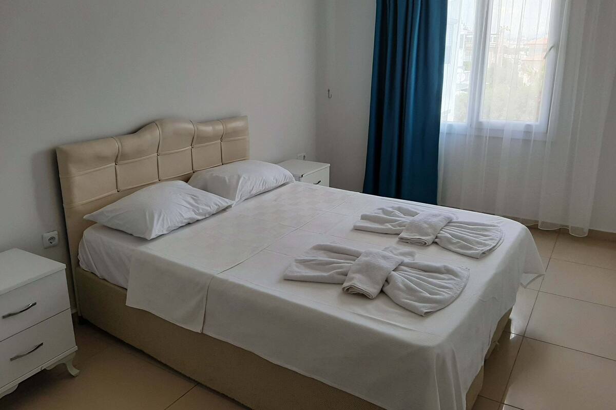 A neatly made bed with white linens and towels, a beige headboard, a white bedside table, and a window with blue curtains.