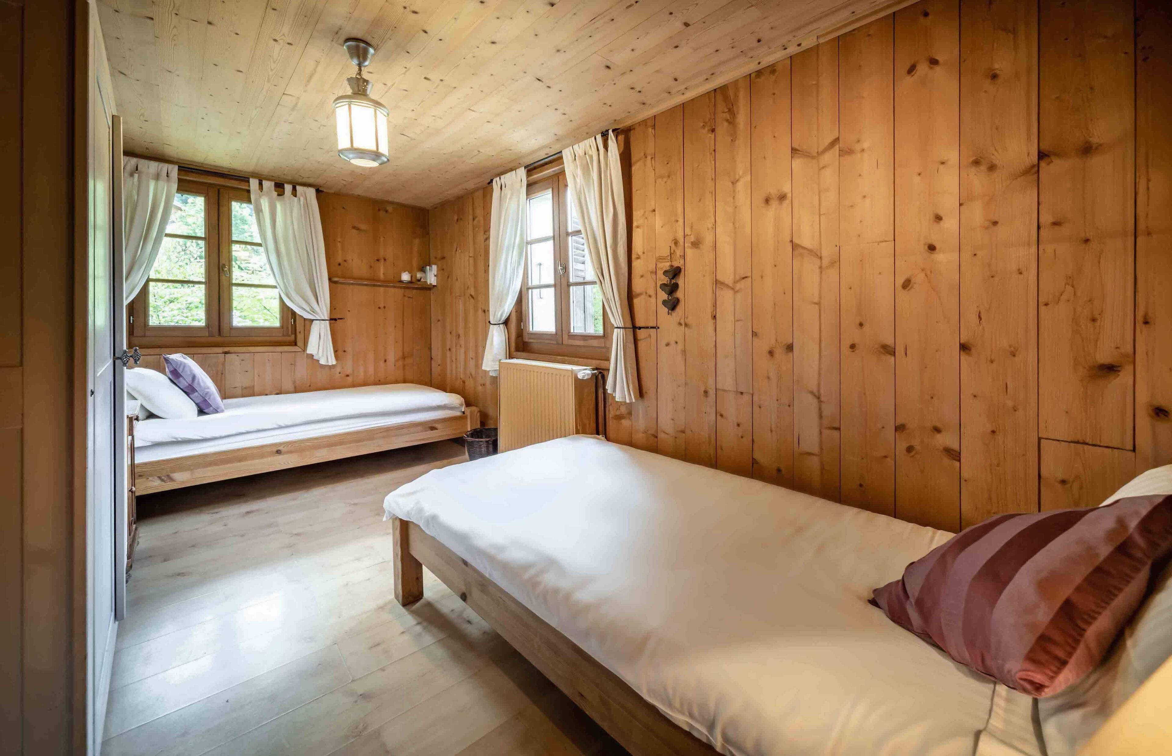A wooden cabin room with two beds, a window with curtains, a radiator, and a ceiling light fixture.