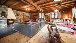 Interior acogedor de una cabaña con chimenea de piedra, sofá de cuero, mesa de comedor de madera dispuesta para una comida y techo rústico de madera.