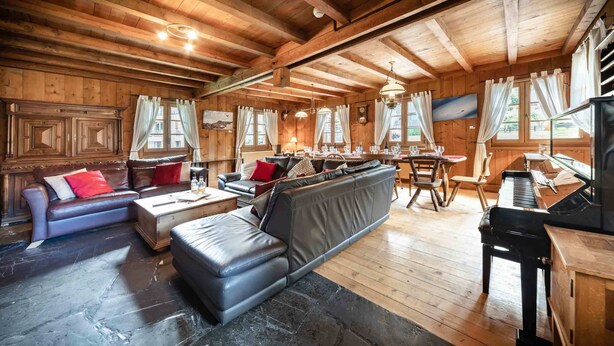 Interior acogedor de una cabaña con suelo de piedra, paredes de madera, un sofá, una mesa de comedor y un piano.