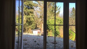 Une pièce dotée d’une grande porte-fenêtre coulissante qui donne sur un espace extérieur agrémenté d’un chemin de gravier, d’arbres et d’un petit abri de jardin.