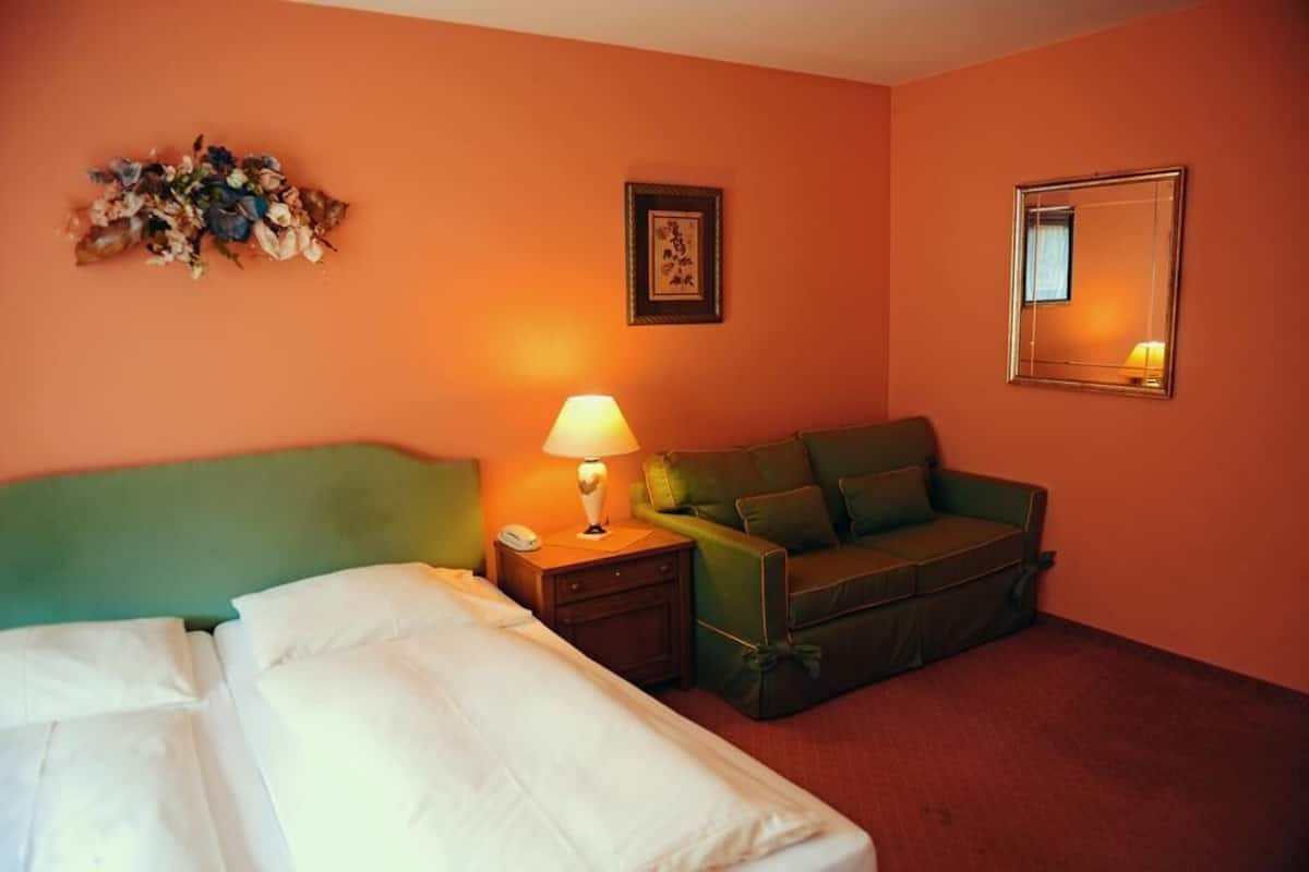 A hotel room with a green headboard, a green sofa, a bedside table with a lamp, and a floral arrangement on the wall.