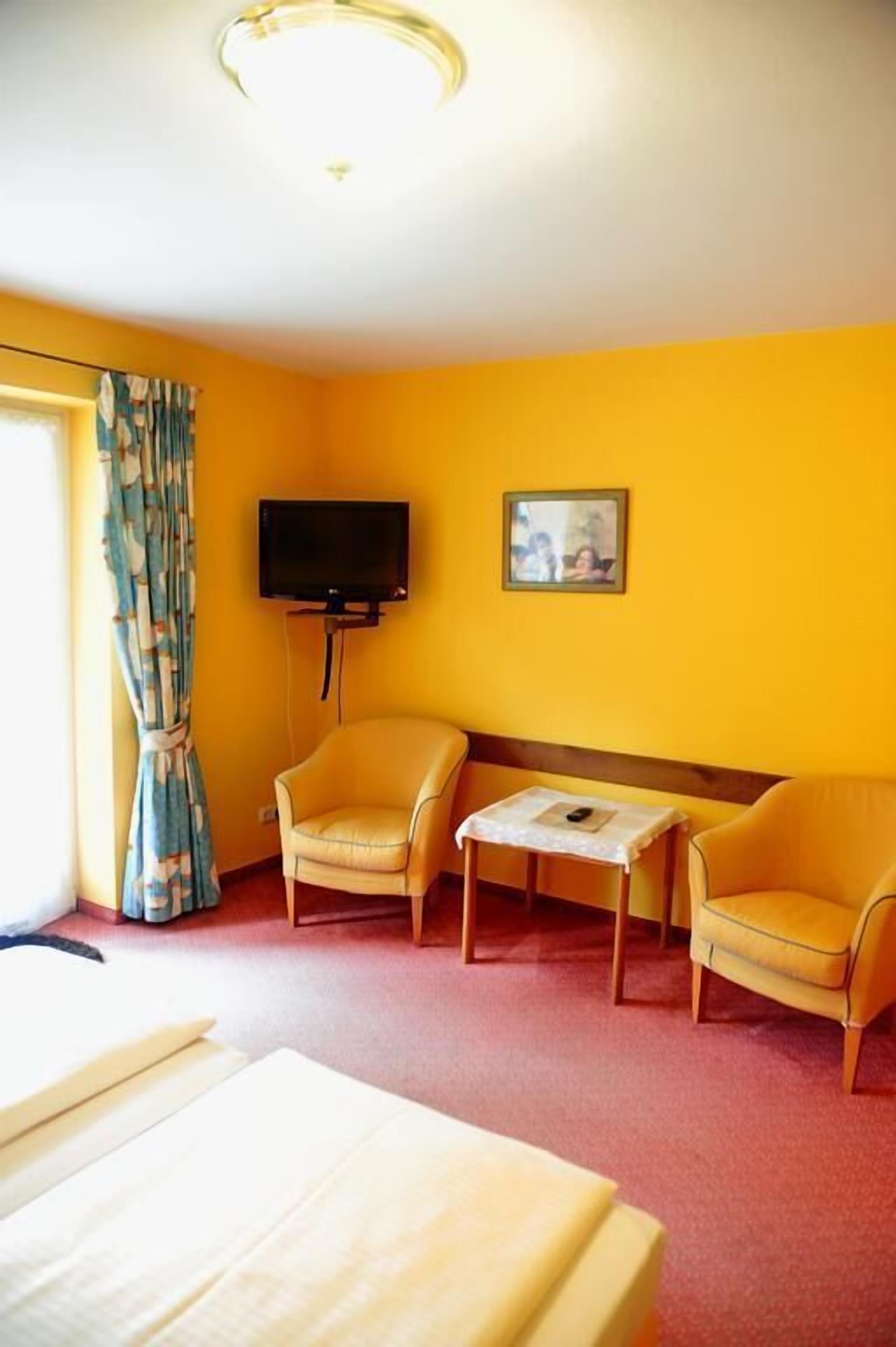 Quarto de hotel com uma cama, duas poltronas, uma mesinha, televisão e janela com cortinas.