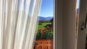 A view through a window with sheer curtains, revealing a wooden deck with red flowers and a green landscape with mountains in the background. - Hotel Kienberger Hof (Pfronten)