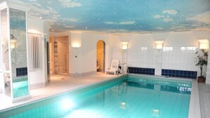An indoor swimming pool with a tiled floor and walls, a ceiling with a cloud design, and a seating area with chairs and a potted plant. - Hotel Kienberger Hof (Pfronten)