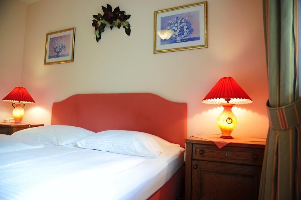 Cama bem arrumada com lençóis brancos, duas luminárias de mesa com abajures vermelhos e duas molduras na parede.