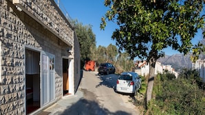 A narrow street with parked cars, a stone building with an open door, and a view of mountains in the distance.